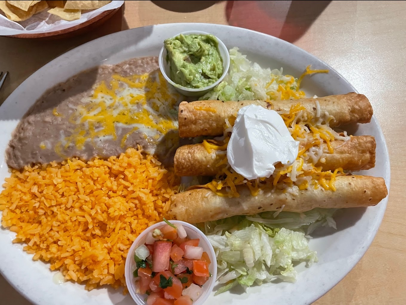 A white plate with three tacos topped with sour cream, a side of rice and beans, lettuce and a dish of Pico de Gallo and guacamole.