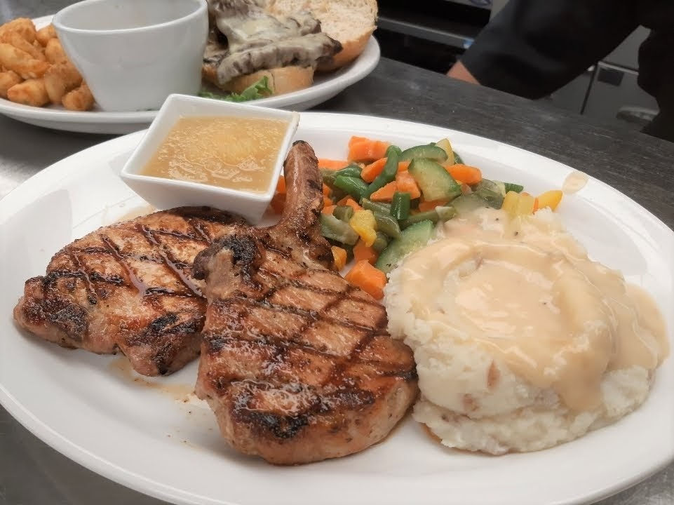 A white plate consisting of a two grilled chicken steak with a side of mashed potatoes and vegetables, with a dish full of app sauce.