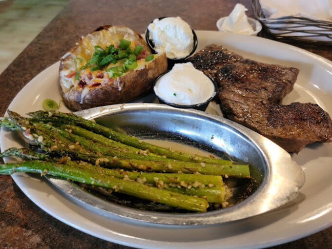 Steak, baked potato, sauce, and a side of asparagus in a metal tin, all on a white plate.