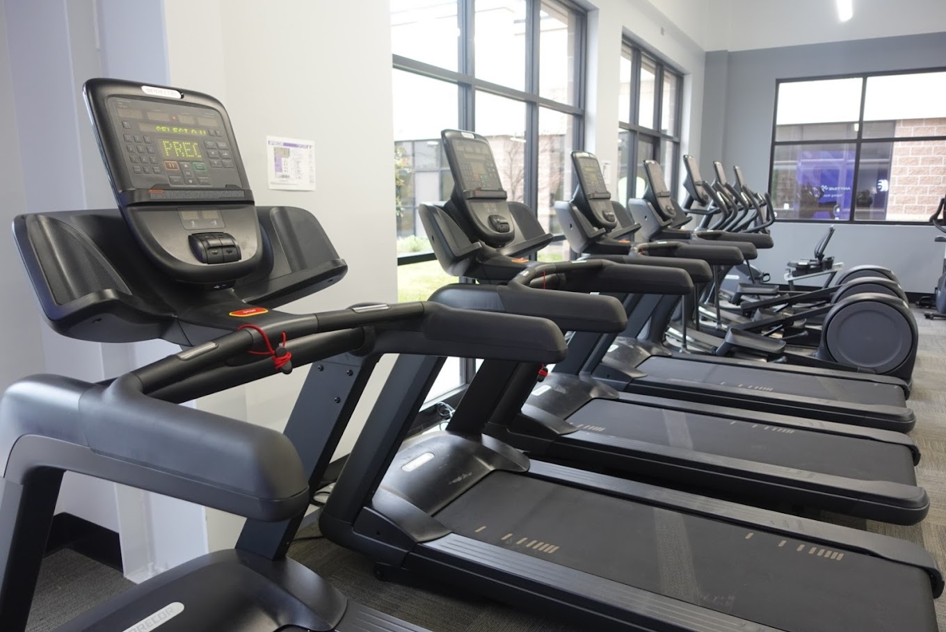 A view of a row of treadmills inside a gym with big windows.