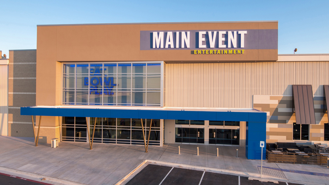 A tan and crème colored building with large windows, and a blue awning over the door. The building reads "Main Event" in white lighted lettering.