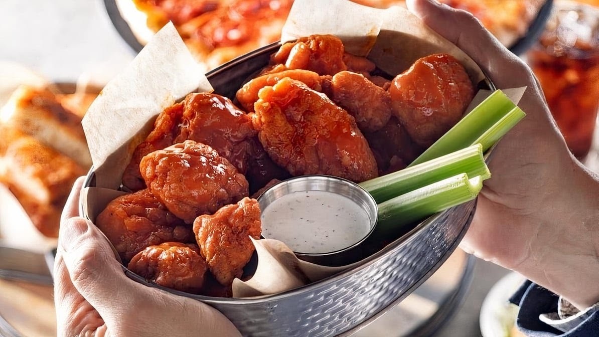 A bowl of wings with a side of ranch and celery, inside of a metal serving bowl and paper to line the bowl.