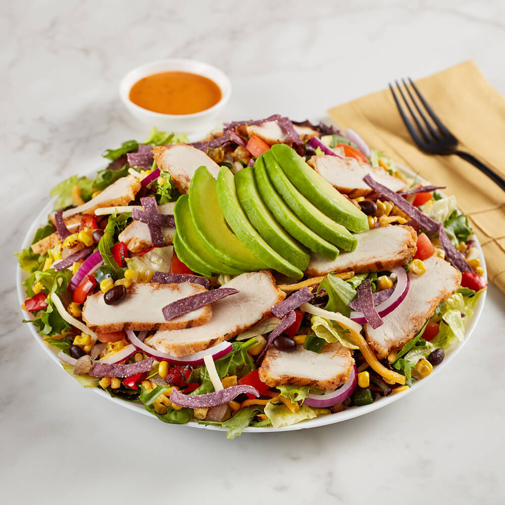 A salad topped with avocado and chicken and a side of sauce.
