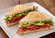 Toasted club sandwich cut into two halves and layered with crispy bacon, lettuce, tomato, avocado, and creamy spread on a white plate.