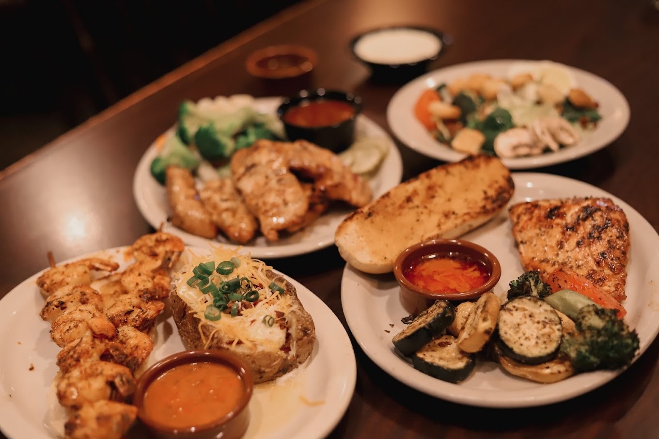 Four white plates full of food and sides from grilled chicken, baked potato, vegetables, and large sides of sauce.