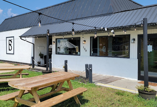 Outside view of a white building with black roof slates. String lights are strung from across the roof. There are two picnic tables on the left side of the image.