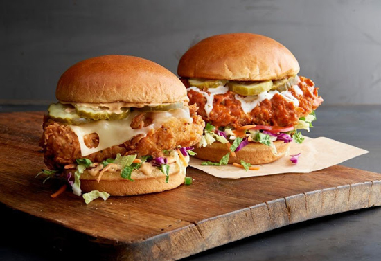 Two burgers with chicken, greens, and cheese set on a wooden plate.