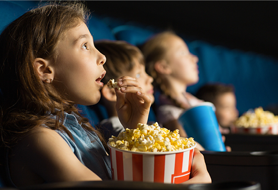 Family of four looking at a movie screen and eating popcorn. Focal point is a girl with her mouth open eating popcorn.