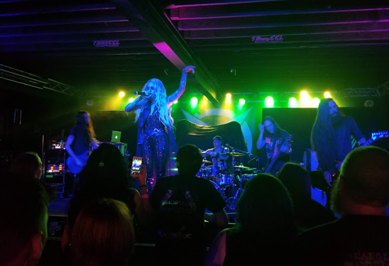 Concert atmosphere with a band on stage and audience members in the foreground. Lighting is yellow, green, and blue.