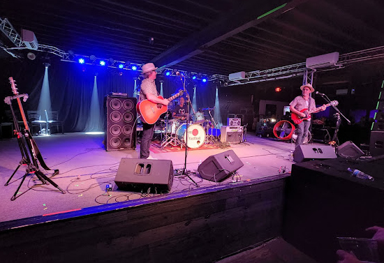 Up close view of stage during a small concert. Two men stand on stage playing guitars in hats.