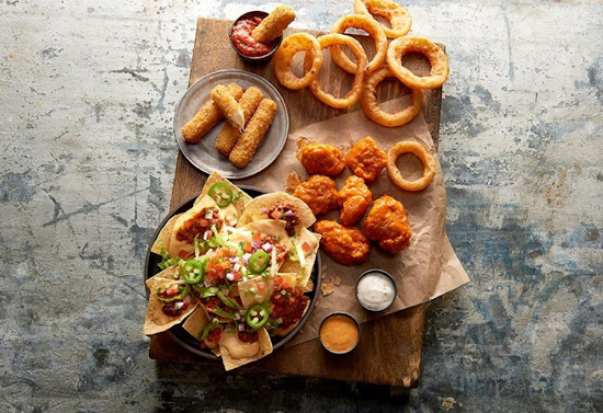 Bowl of chips and sauce, breaded meat, onion rings, and various small sauces on top of a wooden board.