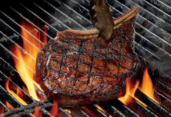 Tongs grabbing a steak surrounded by flames on a grill.
