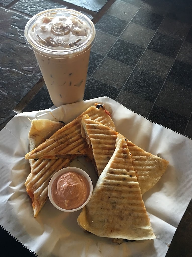 A tray with white serving paper served with a quesadilla a side of sauce and an iced coffee on the side.