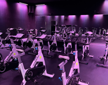 A room light up with dim purple lighting a wall lined with mirrors and a room full of ellipticals.