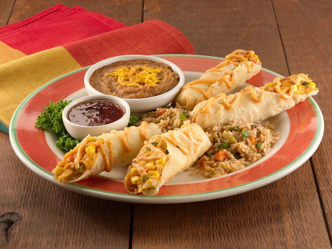 A view of a white plate with a red ring around the edge consisting of four tacos along with a bowl of beans on the side, rice and a small bowl of sauce.