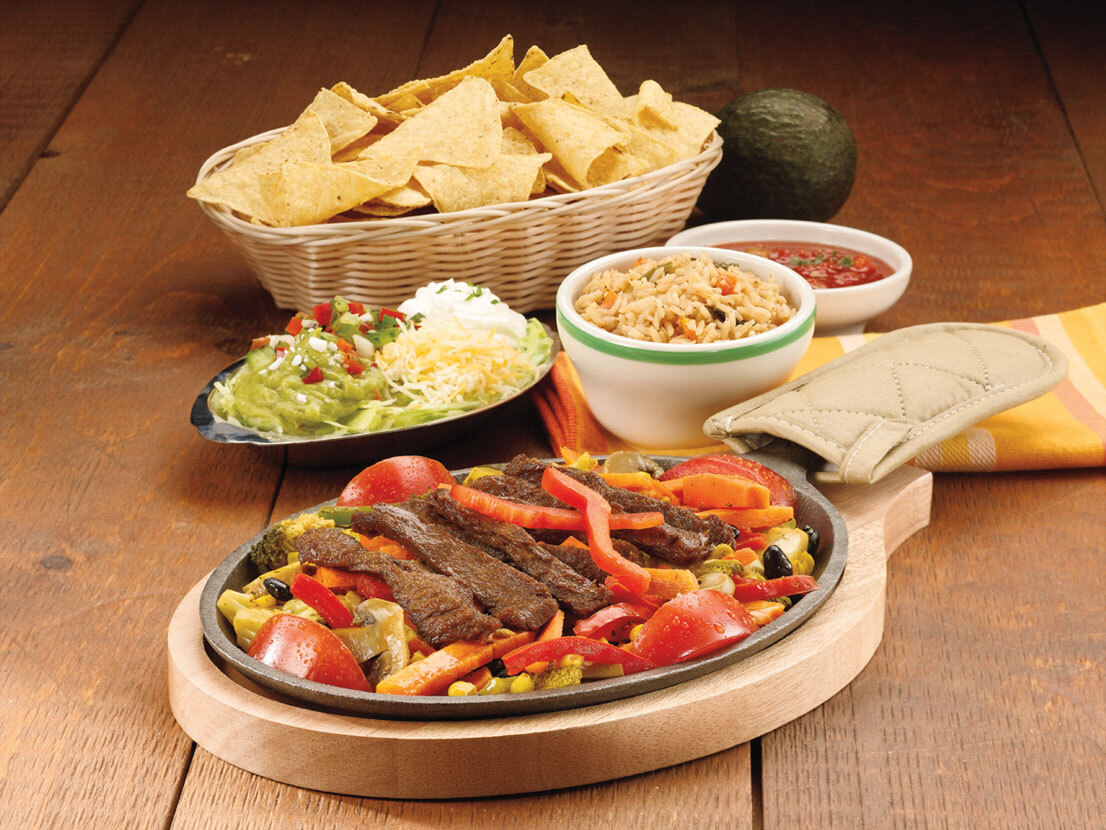 A view of a meal being served on a wooden table, a wooden tray with a large skillet consisting of fajitas, a side of toppings for nachos, and a side of salsa.