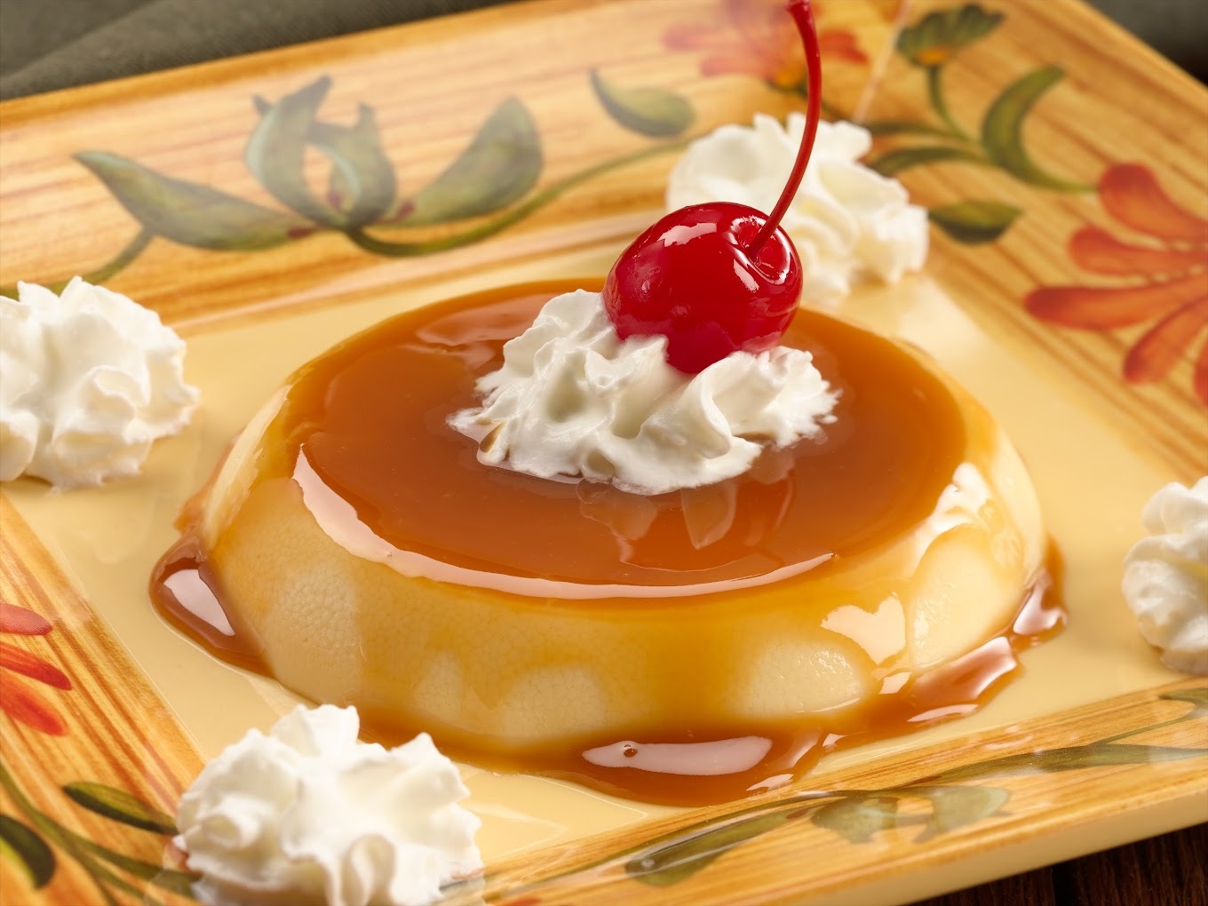 A yellow plate decorated with vines and flowers consisting of a flan desert topped with whipped cream and a cherry.