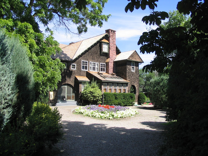 Late 19th century brown brick house with a chimney and shrubbery in front of the entrance. Either side of the photo is framed with green trees.