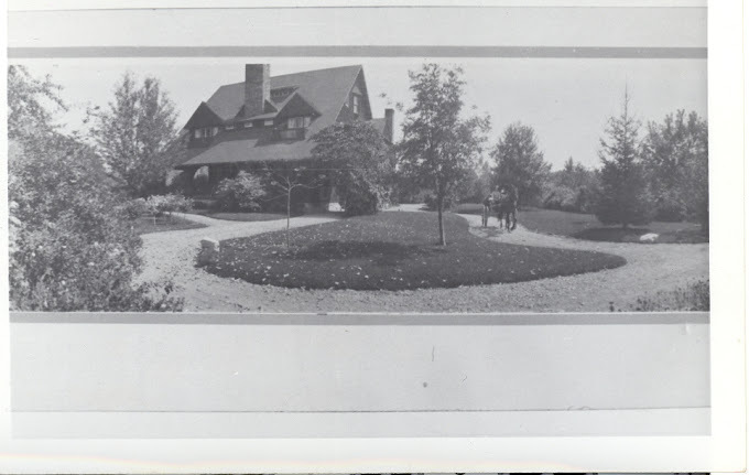 Black and white photo of a large house with a chimney. There is a dirt road and youthful trees surrounding the home.