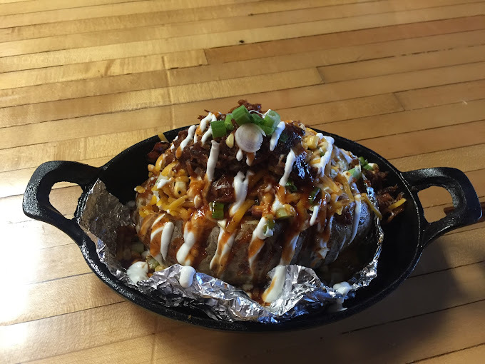 Close view of a foiled baked potato in a cast iron bowl. There are numerous sauces and greens on top of the potato.