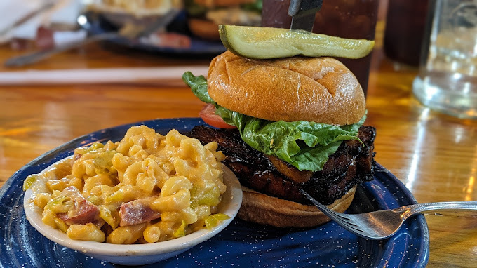 Burger with lettuce and tomato in it, as well as a pickle attached to the top bun. A mac and cheese dish sits in a white bowl, with both food items on the same blue plate.