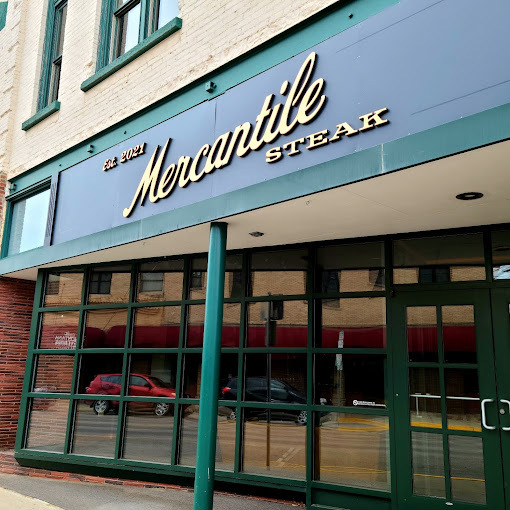 Exterior view of a green and beige building. The windows reflect the road and a small green column is in front of the doors. The sign above the entrance reads "Est. 2021, Mercantile Steak."