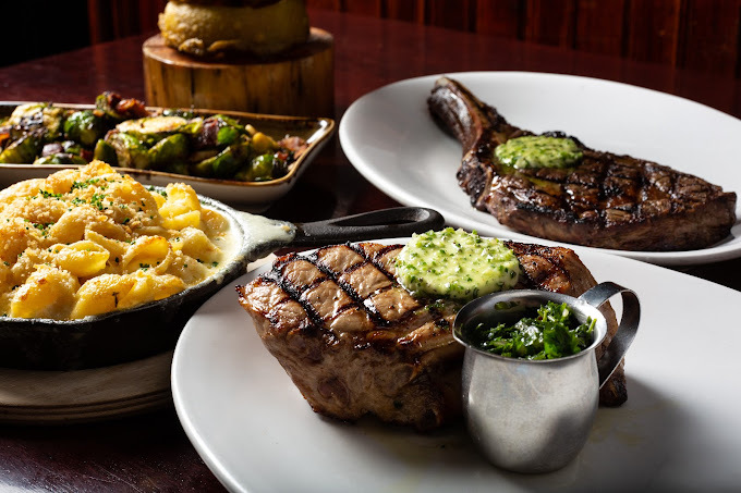 The foreground has a steak with a circular green sauce on it, along with a metal container of greens, on a white plate. The background has another steak on a white plate, a cheesy dish in a cast iron pan, and a dish of greens on a rectangular pan.
