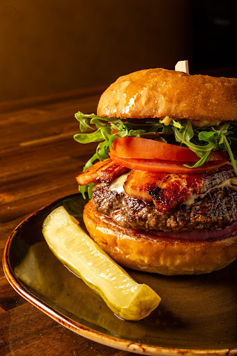 Close view of a burger with tomatoes, lettuce, and bacon, as well as a side of a sliced pickle, on a plate.