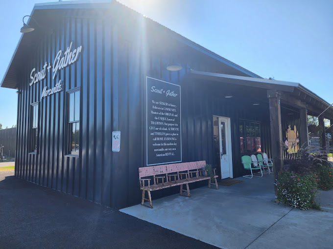 Exterior corner view of a dark vertical slatted building. There is a sun shade above the white door and light green chairs. On the left side of the door, there is a pink wooden bench.