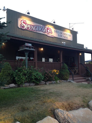 Exterior view of a red building with a front porch and green shrubbery. The sign on the roof reads "Spencer and Co, Steakhouse Lounge." It is lit up by small lights.