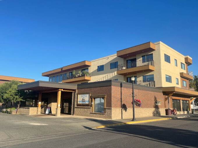 Exterior view of a flat beige building with a flat sun shade. Behind the building is a connected apartment complex.