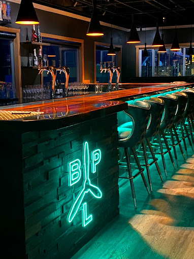 Side view of bar table with teal lighting underneath bar table. Neon sign on the side that has the initials B, P, L, and an image of a fan.