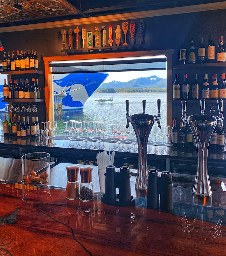 View of bar table with window on the wall. Drinks surround the window. Window view contains the ocean with a large boat.