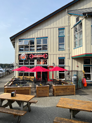 Outside view of a beige slated building. The middle has chairs and tables with red umbrellas and the foreground contains two picnic benches.