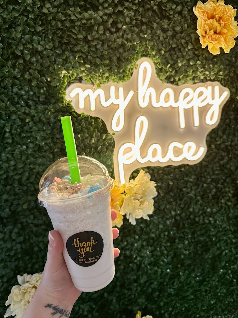 A specialty blended drink topped with Lucky Charms, and displayed behind an greenery wall with flowers and a neon light that reads "My Happy Place" in white light.