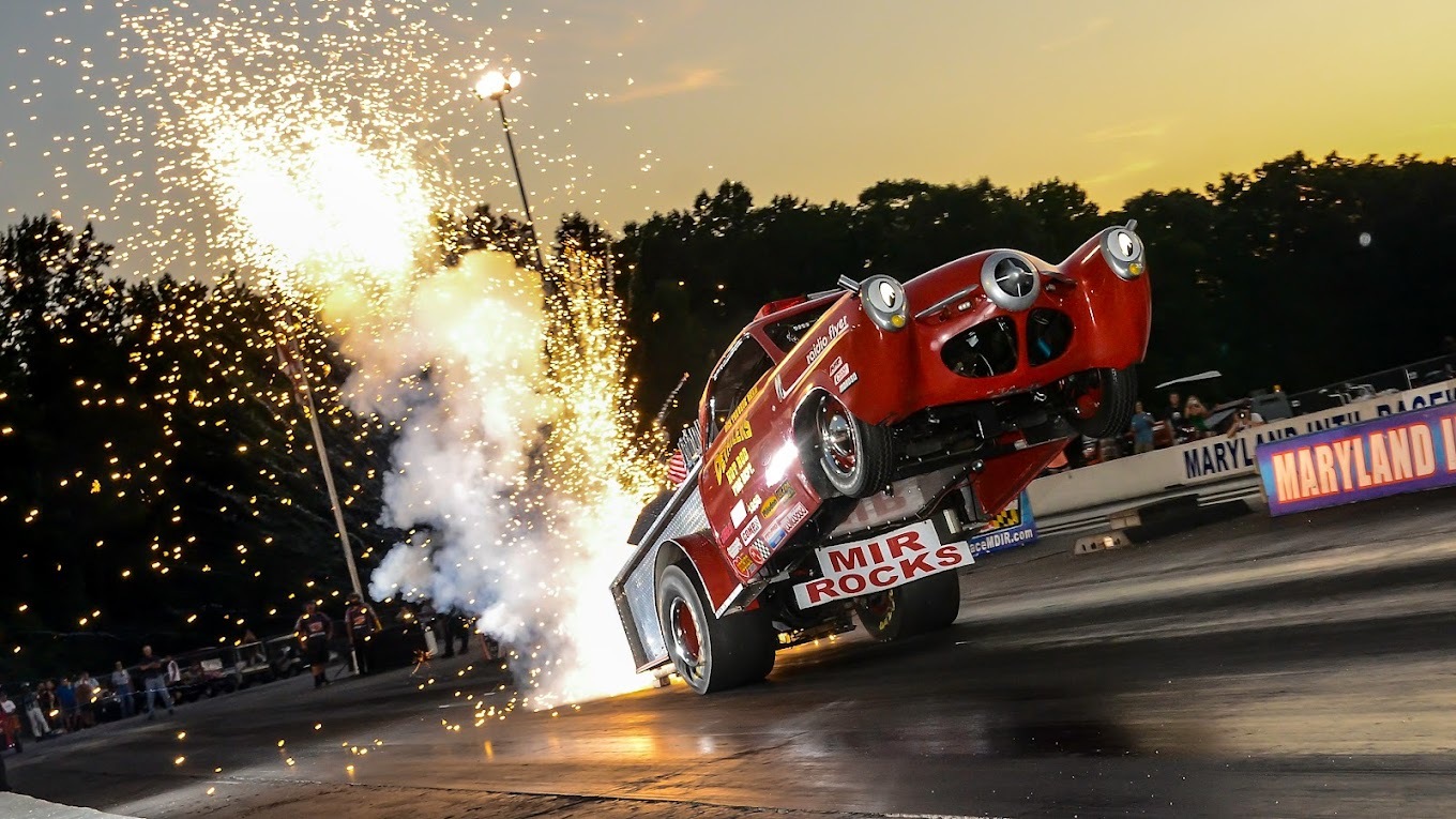 An older vehicle popping a wheelie while fireworks shoot out the back of the vehicle on the drag strip.