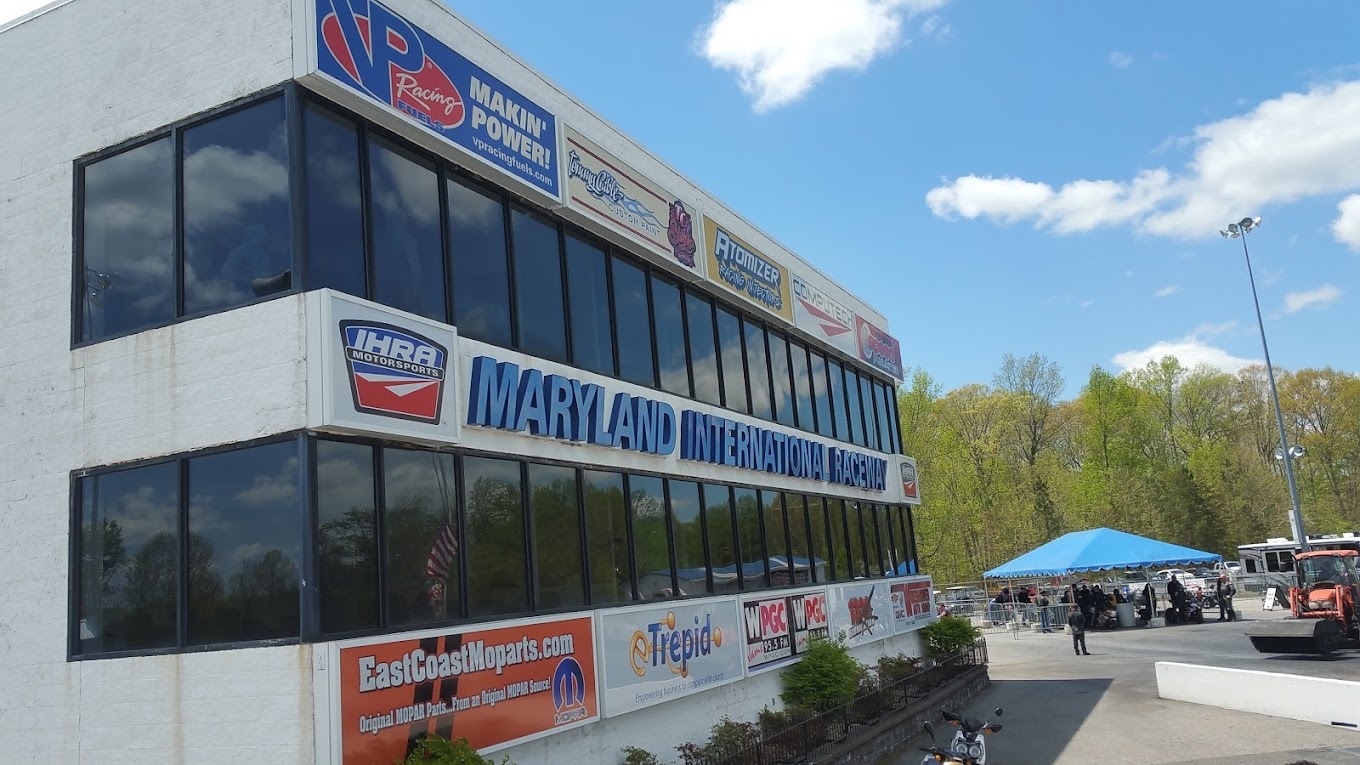 A speedway building with two long strips of windows lining the wall facing towards the track on both floors of the building. Blue lettering lines the front of the building along with sponsorship posters.
