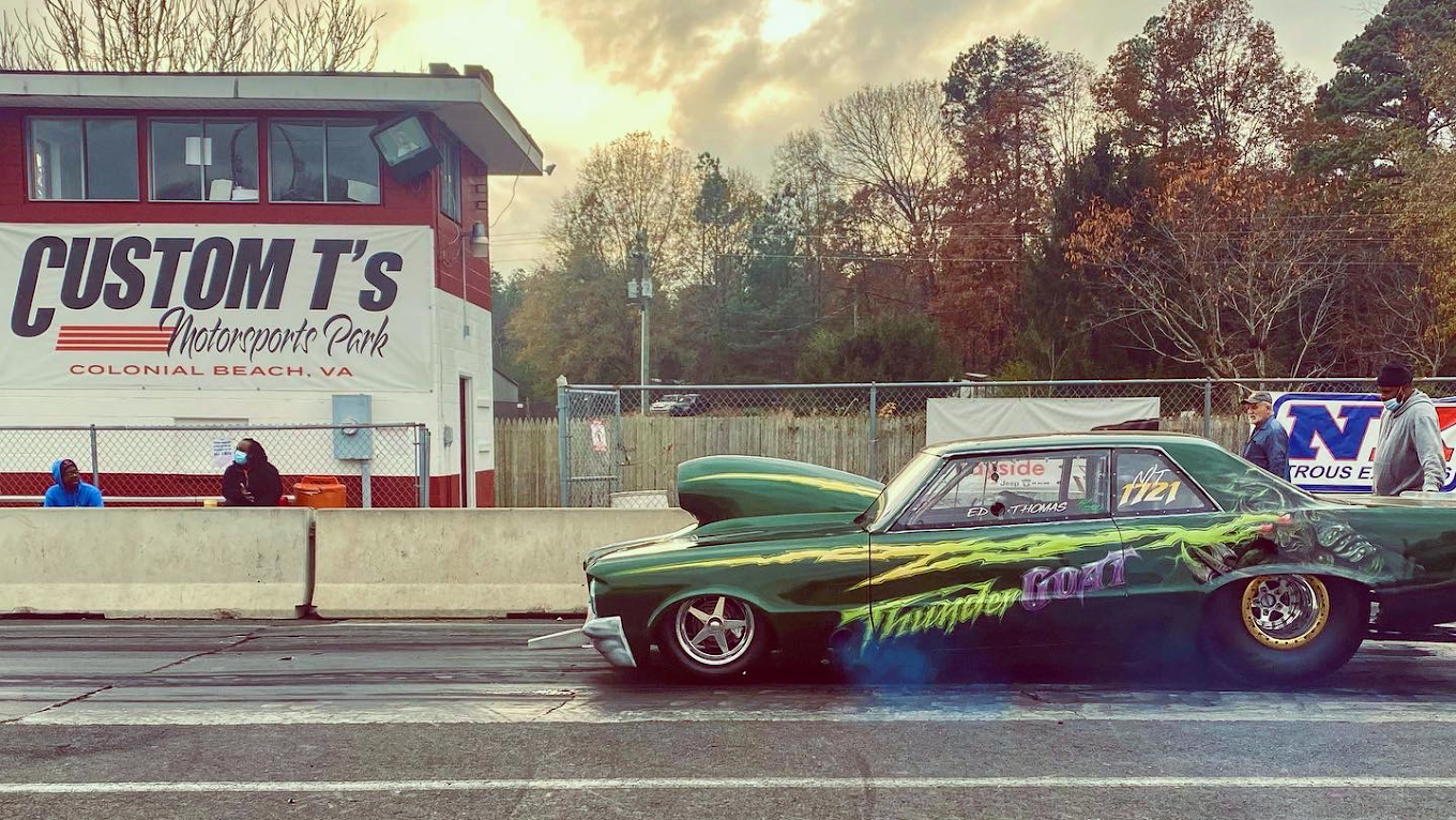 A car with a large engine sits on the drag strip, Custom T's logo on the side of their announcers box in the background behind concrete separators.