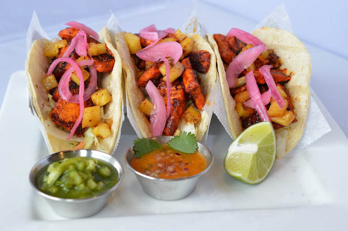 Three small street tacos filled with meat, veggies, and topped with onions along with sliced lime on the side, and two cups of sauce.