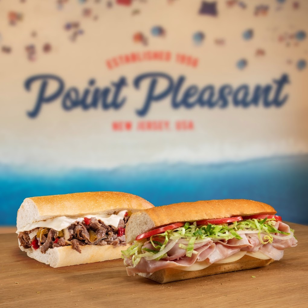 A view of two stuffed cold cut sandwiches sitting on a table with a view of a sandwich shop mural.