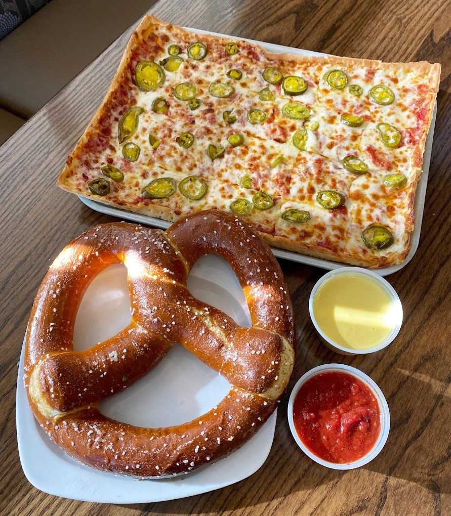 An view of a large rectangular pizza topped with meat cheese and peppers, along with a large soft pretzel on the side with cheese and marinara.