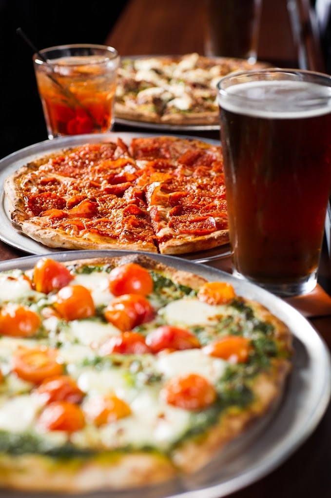 A view of three pizzas with cheese and tomatoes, another with cheese and pepperoni, and another with cheese and chicken. Beers are among the pizzas.