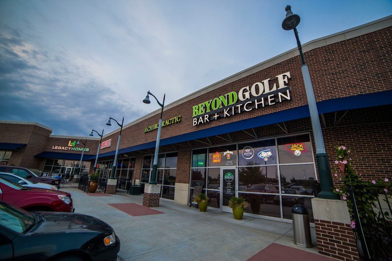 A view of a bar & grill in an strip mall, green and white lettering above the window and door in the center of the space.