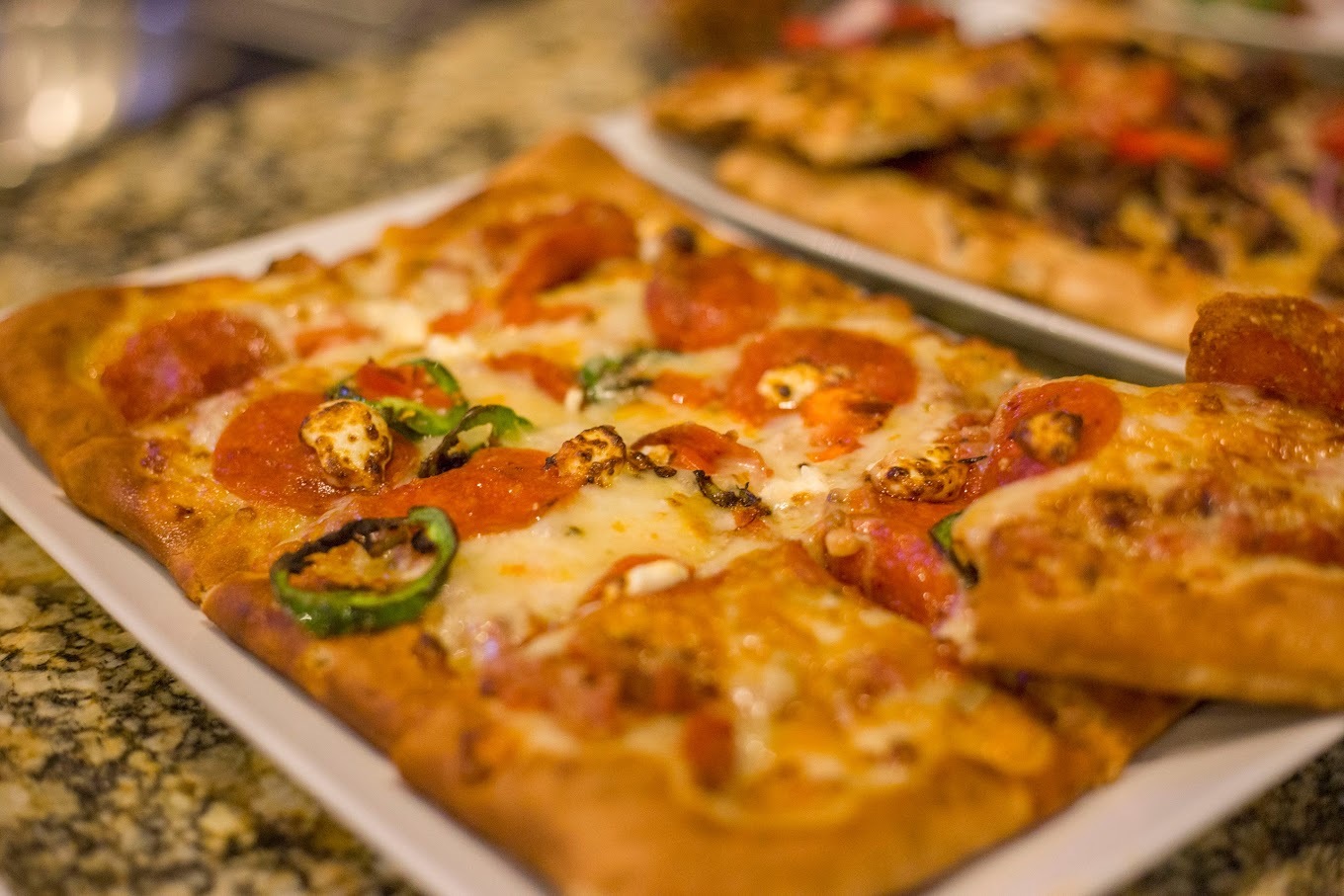 A view of a large rectangular pizza on a white plate, topped with cheese, pepperoni and jalapenos.