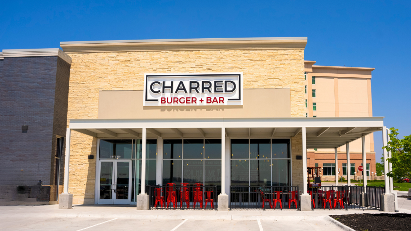 A brick building with an outside seating area with an awning- a large white sign with black and red lettering reading "Charred Burger & Bar"