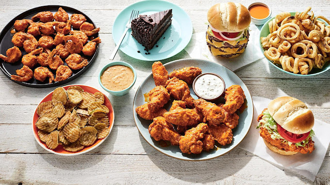 A view of a spread of various entrees such as wings, onion rings, pickle chips, burgers, and chocolate cake.