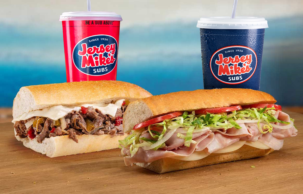 A view of two large cold cut sandwiches with two fountain sodas.