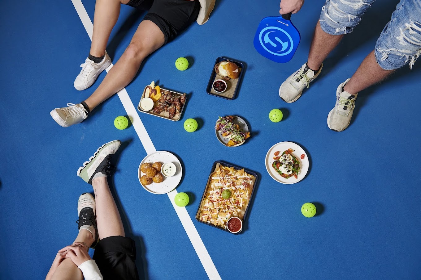 A view of three people sitting on a tennis court surrounded by various dishes such as wings, nachos, and burgers.