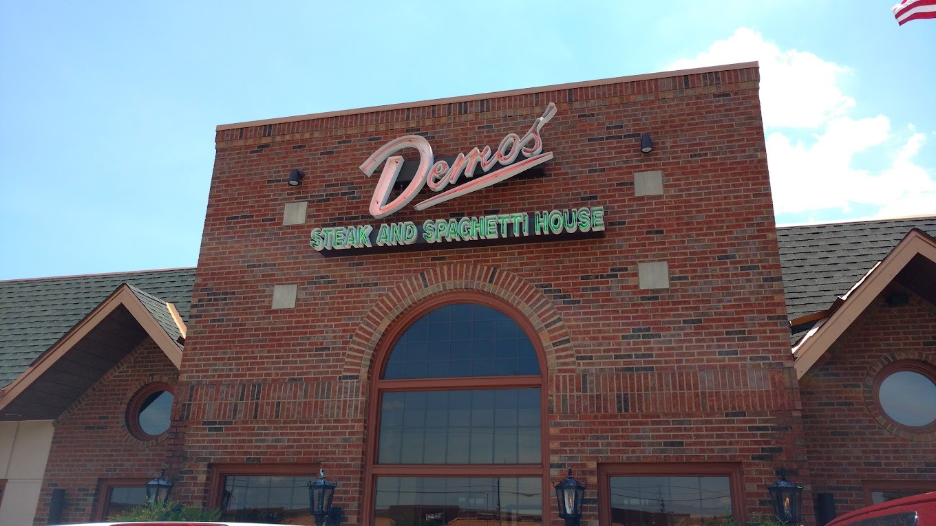 A red brick building with large windows and a large sign reading Demo's in red and Steak and Spaghetti House in green