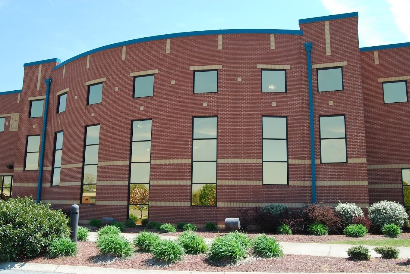A very large red brick building with many windows lining the outside. A sidewalk lined with greenery and shrubs is out front of the building.